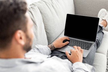 Reklam konsepti. Genç adam boş ekranlı modern dizüstü bilgisayar kullanıyor, koltukta oturuyor, maket yapıyor. Kanepeye yaslanmış milenyum adamı ve boş ekranlı, omuz üstünden vurulan bir defter kullanıyor.