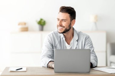 Ofiste dizüstü bilgisayarda çalışan, işyerinde oturan ve boş alana bakan mutlu serbest çalışan bir adam. Başarılı bir iş stratejisi düşünen ya da tatil hayali kuran gülümseyen adam.