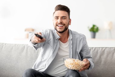 Ev eğlencesi. Neşeli bir adam kanepede dinleniyor uzaktan kumandayla kanal değiştiriyor ve patlamış mısır yiyor, en sevdiği televizyon programını arıyor, evde boş vakit geçiriyor, yer kopyalıyor.