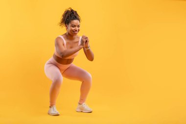 Açık pembe spor kıyafeti giyen bir kadın enerjik bir şekilde canlı sarı bir stüdyoda çömeliyor. Coşku ve kararlılıkla egzersiz yaparken şekline odaklanıyor, boşluğu kopyalıyor.