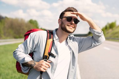 Sırt çantalı yakışıklı genç adam yolda otostop çekiyor, gözleri güneşten koruyor, geçen arabaya bakıyor, kopyalama alanına bakıyor. Otomatik yolculuk, yaz yolculuğu konsepti