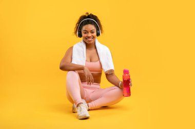 A woman relaxes on the floor after a workout, wearing athletic clothing and headphones. She holds a colorful drink while smiling, surrounded by a vibrant yellow background.