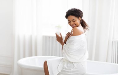 A young African American woman enjoys a relaxing moment in her home bathroom, applying skin care products and feeling beautiful in a serene environment. clipart