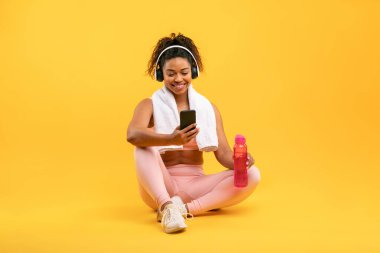 Gülümseyen bir kadın parlak sarı bir arka planda bağdaş kurarak oturur, kulaklık takar ve akıllı telefon tutar. Öte yandan elinde bir su şişesi tutuyor. Egzersiz yaptıktan sonra rahatlamış görünüyor..