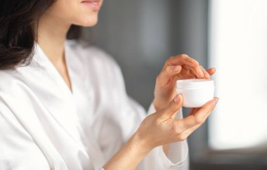 Huzurlu bir ev banyosunda, esmer bir kadın cildini cilt kremiyle şımartmaya odaklanıyor. Elinde küçük bir kavanoz tutuyor. Güzellik rutinini özenle geliştirmeye hazır..