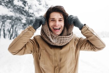 Kış. Başında fular olan yakışıklı bir adam soğuk havada karlı ormanda kameraya gülümsüyor.