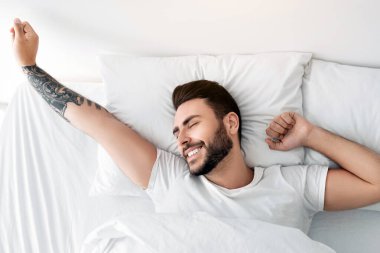 Uyanış konsepti. Mutlu dinlenmiş, iyi uyuyan bir adam sabah uyanır ve ellerini uzatır, kapalı gözlerle gülümserken yatak odasında rahat bir şekilde uzanır, üst manzara