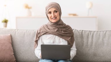 Üst düzey Müslüman kadın dizüstü bilgisayarını kullanarak kameraya gülümsüyor ve başörtüsü takıyor. Oturma odasında bilgisayarlı İslami Kadın pozu. Serbest Kariyer Konsepti