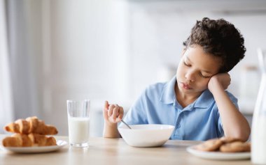 Genç bir çocuk kahvaltı yaparken düşünceli bir şekilde mutfak masasında oturuyor. Kruvasan ve süt de ona eşlik ediyor. Bu da ailesinin yanında sıcak bir aile sabahı geçirdiği anlamına geliyor..