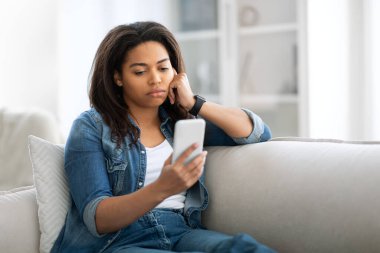 Genç siyahi bir kadın oturma odasında bir kanepede oturur ve dikkatle akıllı telefonuna bakar. Kot ceket giyiyor ve rahat yerinde dinlenirken derin düşüncelere dalıyor..