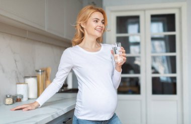 Hamile bir kadın mutfak tezgahının yanında elinde bir bardak su tutarken gülümsüyor. Parlak alan, sakin ve davetkar bir atmosfer yaratan minimalist bir dekor içeriyor..