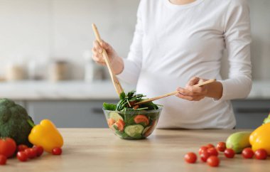 Hamile bir kadın taze sebze salatasıyla yeşillikleri ve renkli dolmalıkları karıştırıyor. Hamilelik boyunca sağlıklı beslenme alışkanlıklarını vurguluyor.