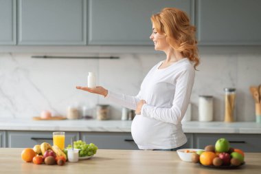 Gülümseyen hamile bir kadın, bir elinde vitamin takviyesi tutarak şık bir mutfakta duruyor. Bu sırada bir dizi taze meyve ve masanın üzerinde bir bardak meyve suyu var..
