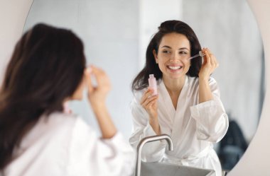 Genç bir esmer kadın yüzüne cilt bakım serumu sürerken banyoda gülümsüyor. Yumuşak bir bornoz giyip özgüven ve güzelliği yansıtan kişisel bakım ritüelini seviyor..