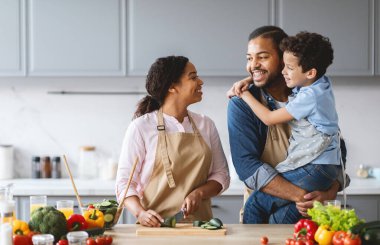 In a bright kitchen, a father holds his son while the mother prepares vegetables. Their smiles reflect happiness and affection as they share a joyful cooking moment as a family. clipart