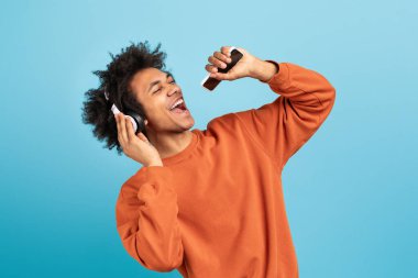 A young African American man expresses joy while listening to music through headphones. He sings along passionately, showcasing a vibrant lifestyle against a blue background. clipart