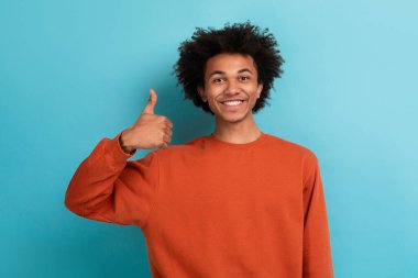 Kıvırcık saçlı genç bir Afrikalı Amerikalı gülümsüyor ve baş parmağını kaldırıyor. Turuncu bir sweatshirt giyiyor, mavi arkaplana karşı canlı bir kontrast oluşturuyor..