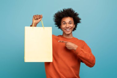 Kıvırcık saçlı neşeli bir Afro-Amerikan genç adam mavi bir arka planda izole edilir. Gülümsüyor ve alışveriş çantasına işaret ediyor..