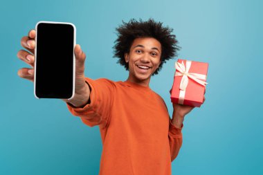 Kıvırcık saçlı genç bir Afro-Amerikalı bir elinde akıllı bir telefon diğer elinde paketlenmiş bir hediye tutarken neşeyle gülümsüyor. Parlak mavi arka plan şenlik havasına katkı yapıyor..