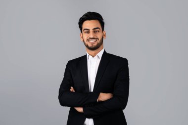 Genç bir Orta Doğulu girişimci stüdyoda kollarını kavuşturmuş dururken kendinden emin bir şekilde gülümsüyor. Mesleki kıyafetleri, başarı ve yönetime odaklanmış kurumsal bir yaşam tarzını yansıtıyor..