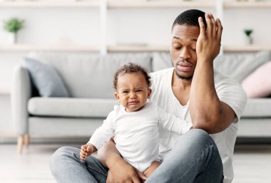 Yorgun, genç bir baba evde yerde ağlayan yeni doğmuş bebekle oturuyor, umutsuz Afrikalı Amerikalı baba bebeğini üzüyor, babalık izni sırasında acı çekiyor.