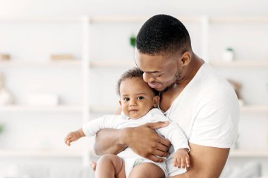 Baba ve Çocuk. Mutlu Afrikalı Amerikalı bir babanın portresi, ellerinde şirin bir erkek bebek, iç mekanda birlikte poz veriyor, sevimli siyah çocuk kameraya bakıyor ve gülümsüyor, boşluğu kopyalıyor.