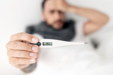 Hasta genç adam kameraya termometreyi gösteriyor, yatağa uzanıyor ve alnına dokunuyor. Coronavirus, grip belirtileri. Seçici odak, üst görünüm