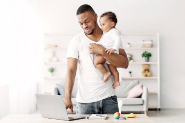 Ellerinde küçük bir bebek tutan ve dizüstü bilgisayar kullanan genç siyahi adam, Millennial Afrikalı Amerikalı baba, sevimli bebeğine bakıcılık yaparken bilgisayarla uzaktan çalışıyor.