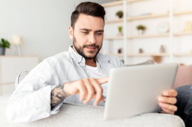 Evdeki iç dekorasyonu kullanarak kanepeye yaslanmış yakışıklı bir adamın yakın çekimi. Oturma odasındaki koltukta dinlenen mutlu adam dijital tablet, fotokopi alanı ve panorama üzerinde mobil uygulama kullanıyor.