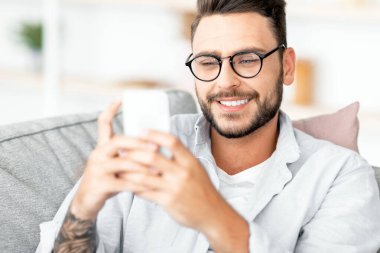 Ev içi rahatlık. Akıllı telefon kullanan, oturma odasındaki koltukta dinlenen, cep telefonuyla mesajlaşırken gülümseyen, rahat bir koltukta oturan mutlu bir adam.