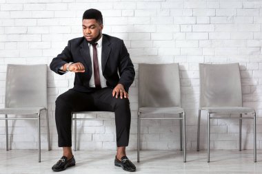 Resmi giyinmiş genç bir Afro-Amerikalı saatine bakıyor, saati kontrol ediyor, ofis lobisinde iş görüşmesini bekliyor, fotokopi odasında. Personel alımı kavramı