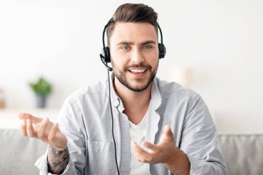 Kulaklıklı heyecanlı bir adam web kamerasıyla konuşuyor uzaktan çalışan bir meslektaşla video görüşmesi yapıyor, evdeki kanepede oturuyor. Video konferansı, modern iletişim, çevrimiçi iş eğitimi kavramı