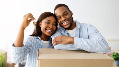 Emlak konsepti. Afro-Amerikalı genç eşleri sevmek taşınan kutular ve yeni ev anahtarlarıyla poz vermek, kameraya sarılmak ve gülümsemek, kutularla dolu bir odada durmak.
