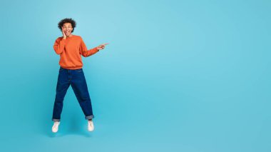Kıvırcık saçlı genç bir Afrikalı Amerikalı neşeli bir şekilde atlıyor, parlak bir süveter ve rahat bir kot pantolon giyiyor, fotokopi alanını işaret ediyor.
