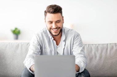 İşe alım konsepti. Mutlu milenyum adamı internette iş fırsatlarını araştırıyor, iş arama uygulamasını ya da internet sitesini kullanıyor, evdeki kanepede oturuyor, boş yer arıyor.