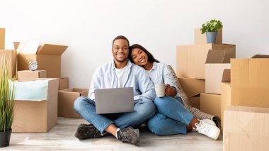 Taşınma konsepti. Mutlu Afro-Amerikalı çift yeni daire için mobilya alıyor, yerde oturuyor ve dizüstü bilgisayar kullanıyor, boş yer var. Heyecanlı eşler kameraya gülümsüyor, kutuların arasında oturuyor.