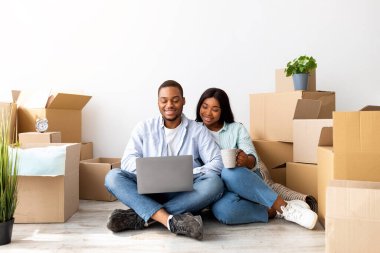 Mutlu Afro-Amerikan eşleri yeni eve taşındıktan sonra dizüstü bilgisayarlarıyla kutuların arasında oturuyorlar, internette geziniyorlar, boş alanlar var. Gayrimenkul, ev sahipliği ve taşınma kavramı