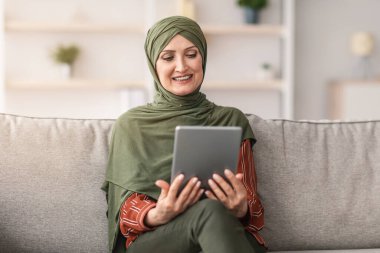 Mutlu orta yaşlı Müslüman kadın tesettürde otururken Tablet Bilgisayar kullanarak online film seyrediyor. Modern Orta Doğulu Kadın İnternet Taraması, Dijital Gadget Kullanımı