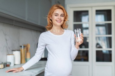 Gülümseyen hamile bir kadın parlak bir mutfakta elinde temiz bir bardak suyla duruyor. Basit, uzun kollu bir gömlek giyiyor. Evinde sağlık ve mutluluk hissi yayıyor..