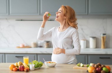 Gülümseyen hamile bir kadın şık bir mutfakta durmuş taze meyve suyunu yudumlarken neşeyle renkli meyve salatası yiyor. Sahne çeşitli meyveler ve bir bardak portakal suyuyla dolu..