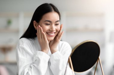 Bornozlu güzel bir Asyalı kadın yüz masajı yapıyor, yatak odasında masada oturuyor, aynaya bakıyor ve gülümsüyor, güzel Çinli kadın duştan sonra nemlendirici yüz kremi kullanıyor, boşluğu kopyalıyor.