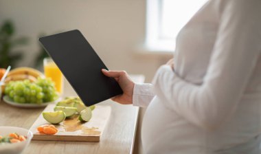 Hamile bir kadın, besleyici yemekler planlarken elinde tabletle parlak bir mutfakta duruyor. Taze meyve ve sebzeler tezgahta sergileniyor, sağlıklı bir yaşam tarzını teşvik ediyor, kırpılıyor.