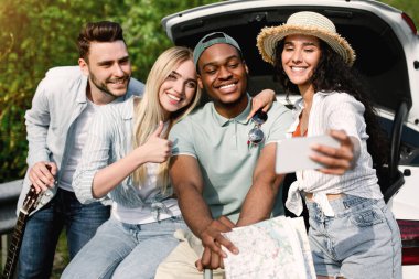 Yolculuk sırasında cep telefonuyla selfie çeken, bagajda oturan, yazın birlikte seyahat eden tuhaf genç arkadaşlar. Çok uluslu milenyum insanları yol kenarında eğleniyor, fotoğraf çekiyorlar.
