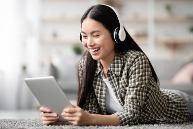 Güzel Asyalı kadın film izliyor, evde yerde yatarken kablosuz kulaklık ve ped kullanıyor, yakın plan, kopyalama alanı. Mutlu genç kadın dijital tablette eğlenceli yeni mobil uygulamalarla eğleniyor