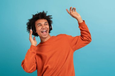 Kıvırcık saçlı genç bir Afrikalı Amerikalı kulaklıkla müzik dinlerken neşeyle dans ediyor. Mutluluk ve enerjiyi canlı mavi bir ortamda ifade ediyor..