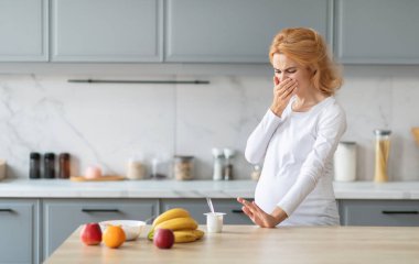 A pregnant woman stands in her stylish kitchen, covering her mouth in discomfort. Fresh fruits like bananas, apples, and oranges are on the counter, alongside yogurt. clipart