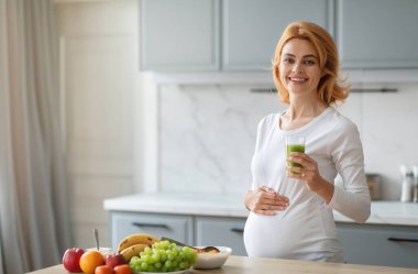 Hamile bir kadın modern bir mutfakta elinde bir bardak yeşil smoothie ile duruyor. Taze meyveler, elmalar, muzlar ve üzümler tezgahın üzerine yerleştirilmiş, kopyalama alanı