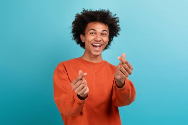 Kıvırcık saçlı genç bir Afrikalı Amerikalı gülümseyerek ve neşeli el hareketleri yaparak neşe ifade ediyor. Parlak mavi bir arka plana karşı duruyor, pozitif enerji yayıyor..
