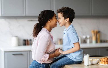 Mutfakta bir anne ve oğlunun kahvaltı ederken şefkatli bir gülümseme paylaştıkları neşeli bir an. Şefkatli etkileşimleri aile sevgisini ve ebeveynliği yansıtır..