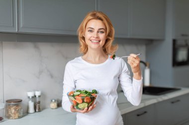 Elinde bir kase salata tutarken gülümseyen sarı saçlı hamile bir kadın. Çatalla ısırır, doğal ışıkla dolu şık bir mutfakta canlı sebzeler sergiler..